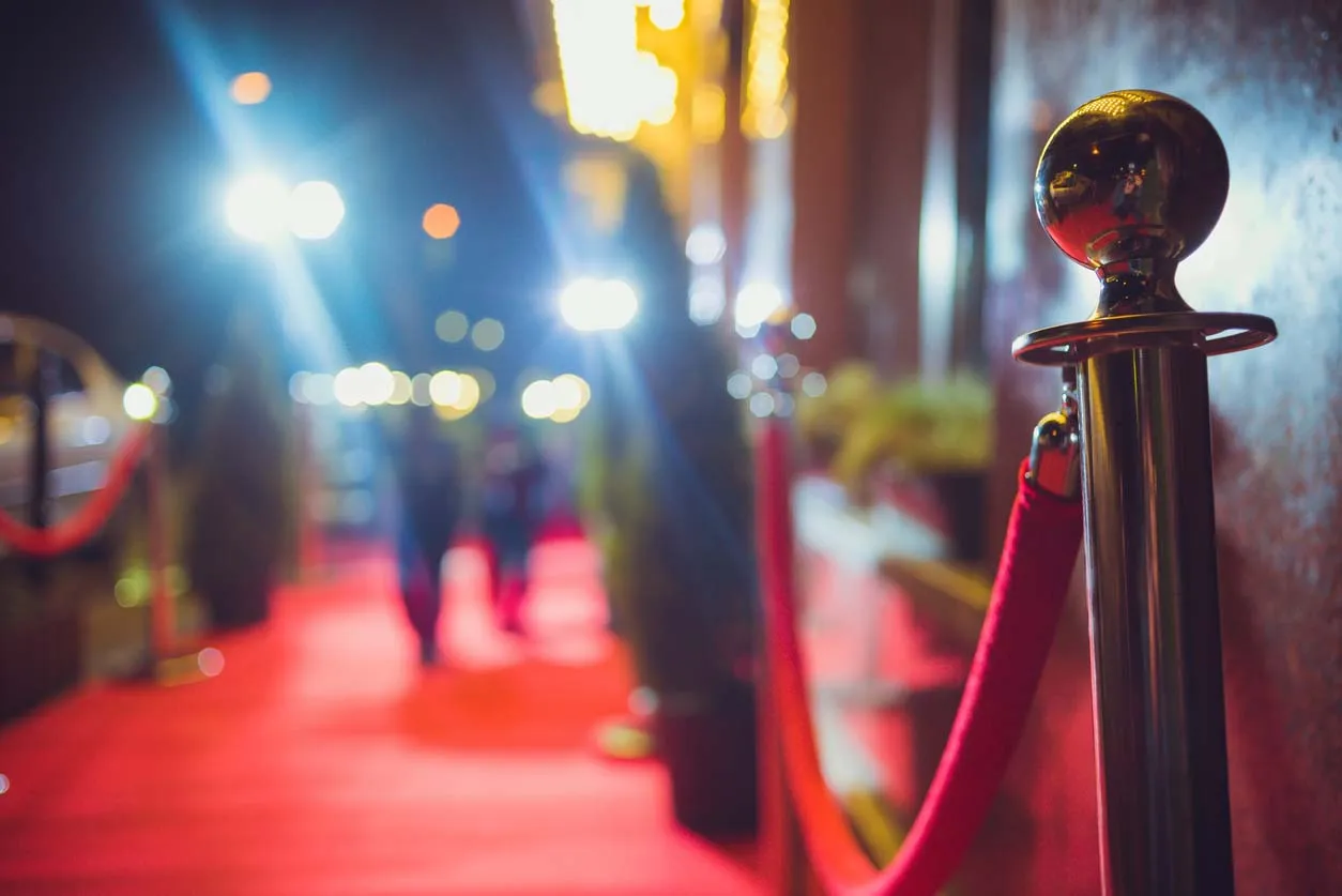 Vue floue d'un tapis rouge illuminé la nuit, avec une barrière en corde rouge et un poteau métallique brillant au premier plan, créant une ambiance d'événement prestigieux.